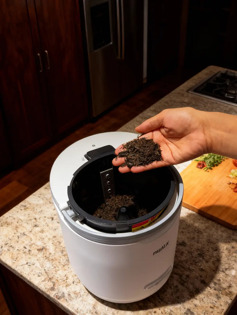 A real-life demonstration of the Mb4 smart composter on a kitchen countertop, with a hand holding a handful of fresh, dark organic fertilizer produced from daily food waste.
